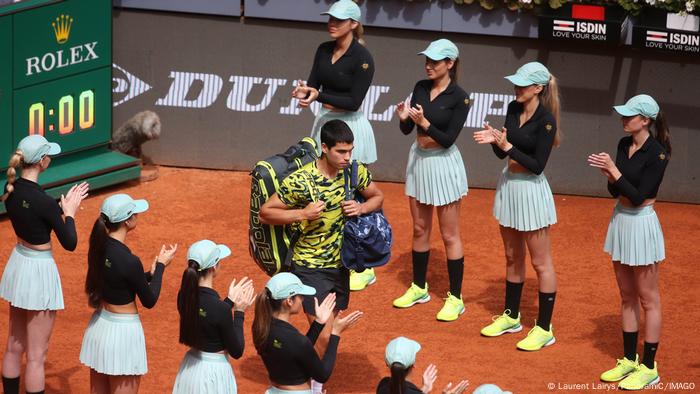 El jugador de tenis Carlos Alcaraz pasea entre dos filas de recogepelotas modelos, vestidas con falda plisada corta y tops que dejan ver el ombligo. En la cabeza, llevan una gorra.