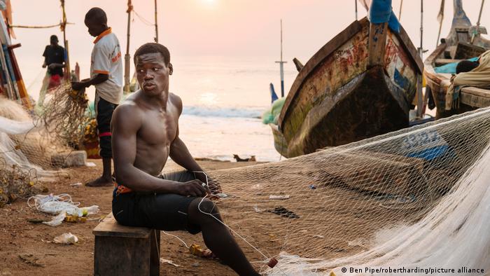 Ein Mann sitzt vor einem aufgespannten Fischernetz; im Hintergrund sind weitere Menschen und hölzerne Kanus zu sehen, die in Sicherheit vor der Brandung des Atlantiks liegen.