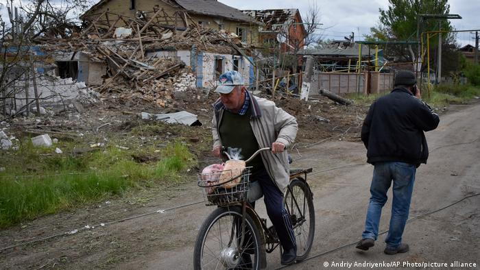 Ein Mann auf einem Fahrrad und ein Mann, der mit einem Mobiltelefon telefoniert, stehen auf einer unbefestigten Straße, im Hintergrund ist ein zerstörtes Hasu zu sehen, Region Saporischschja in der Ukraine