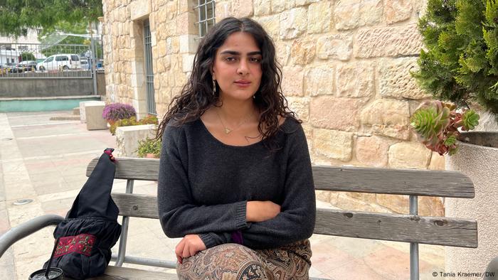 Faizeh Afifi sitzt in schwarzem Pullover und gemustertem Rock auf einer öffentlichen Bank und blickt ernst in die Kamera Faizeh Afifi sitzt in schwarzem Pullover und gemustertem Rock auf einer öffentlichen Bank und blickt ernst in die Kamera