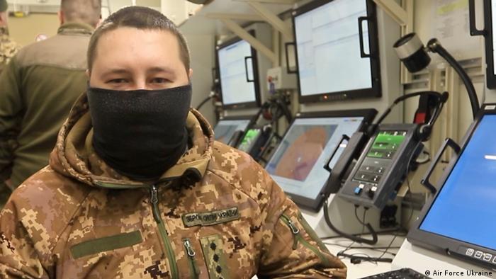 Ukrainischer Soldat in Uniform mit schwarzer Maske bis unter die Augen im Kontrollstand des Iris-T SLM Flugabwehrsystem, im Hintergrund Monitore und Steuerungsgeräte