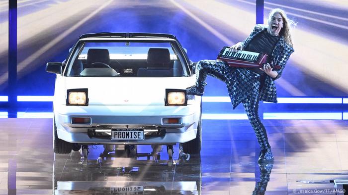Ein Auto steht auf einer Bühne, daneben posiert ein Musiker mit einem tragbaren Keyboard.