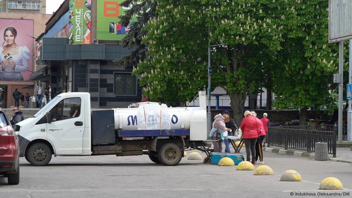 A milk truck is parked on a street.  A woman sells milk at a small table right next to the milk truck