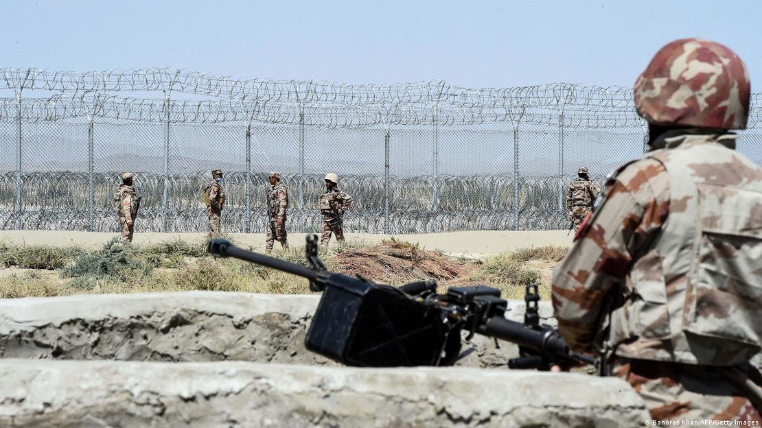Un soldado junto a un fusil automático observa a otros soldados que patrullan junto a una valla fronteriza.