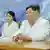 North Korean leader Kim Jong Un and his daughter Kim Ju Ae sit at a long table, against a blue and white background. Both are dressed in white