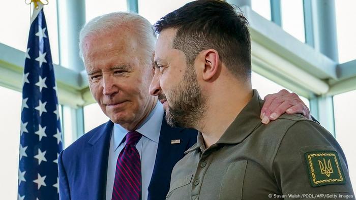 Biden legt seinen Arm um Selenskyj, beide lächeln, neben ihnen eine amerikanische Flagge