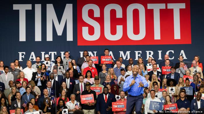 El candidato sonríe con el micro en la mano y un grupo de gente detrás, con un gran cartel con su nombre al fondo en el que se lee fe en América.