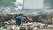 Se ve al niño agachado recogiendo algo y mirando a cámara, con un camión abandonado al fondo y basura desperdigada. Se ve al niño agachado recogiendo algo y mirando a cámara, con un camión abandonado al fondo y basura desperdigada.