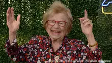 Eine ältere Frau in geblümter Bluse und mit rot gerahmter Brille hebt die Hände und lächelt in die Kamera.