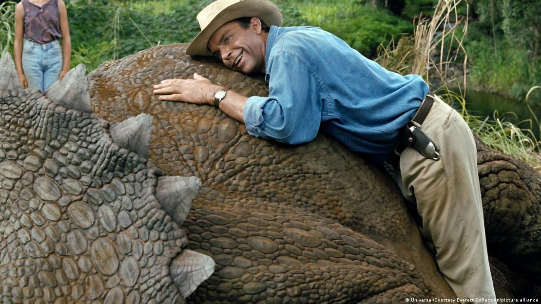 Homem sorri ao se debruçar no dorso de um dinossauro