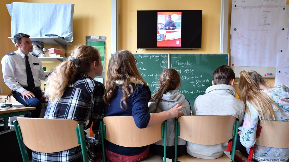 Ein Polizist und mehrere Schulkinder verfolgen an einem an der Wand hängenden Bildschirm einen Beitrag 