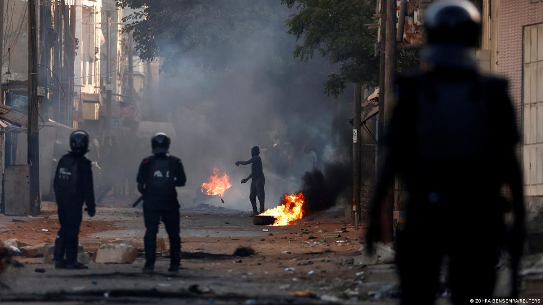 A thick cloud of smoke rises from smaller fires along the street, with a protester and three police officials in the picture, as violent crashes break out in Dakar in early June following Sonko's conviction
