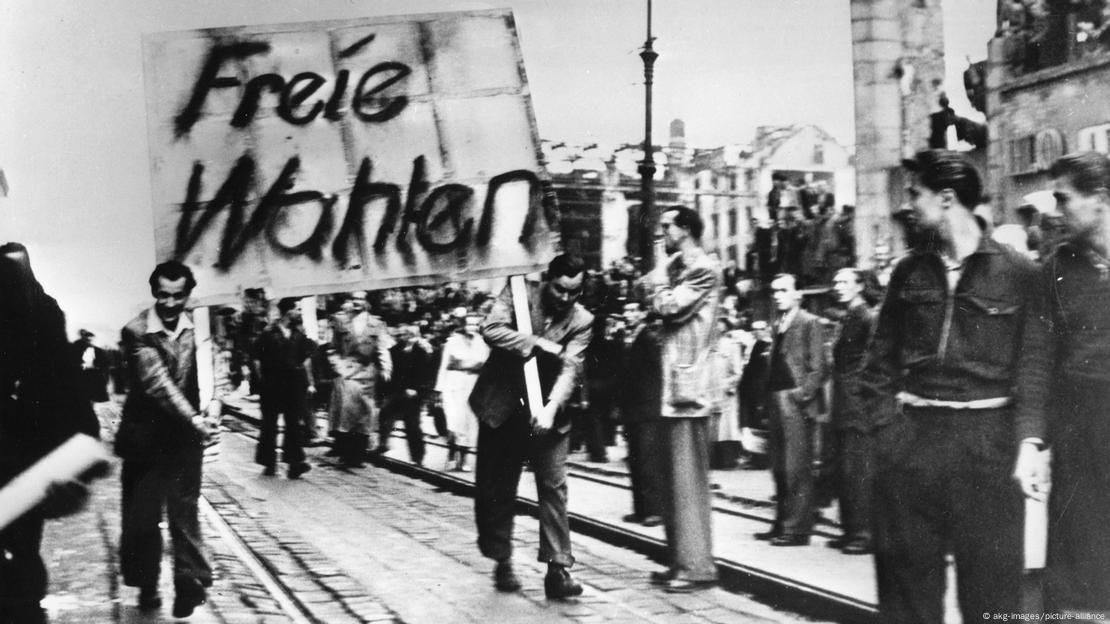 Dos personas sostienen una improvisada pancarta en la que se lee "elecciones libres" mientras la multitud los mira pasar. Dos personas sostienen una improvisada pancarta en la que se lee "elecciones libres" mientras la multitud los mira pasar.