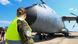 A female soldier in uniform and yellow high-visibility vest stands in front of a military transport plane. A female soldier in uniform and yellow high-visibility vest stands in front of a military transport plane.
