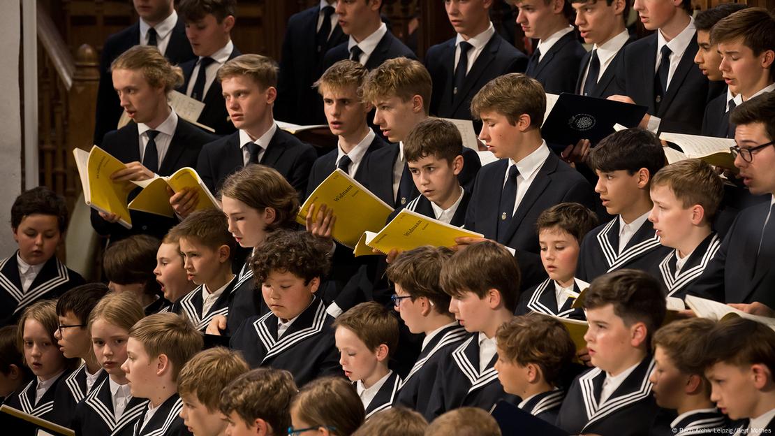 Picture of an all boys choicr where all choir members are dressed in black and white and are holding their sheet music that has yellow front and back covers.