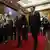 Two men dressed in suits walk down a corridor on a red carpet, flanked by people taking photos
