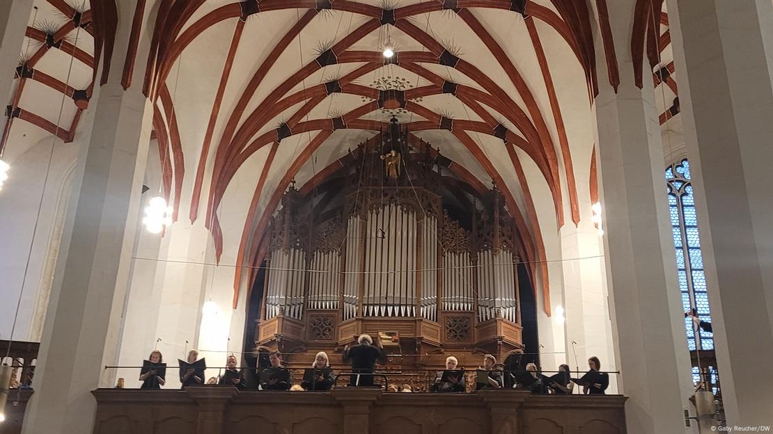 Picture of a man conducting a choir and orchestra who are performing on the balcony of a church.