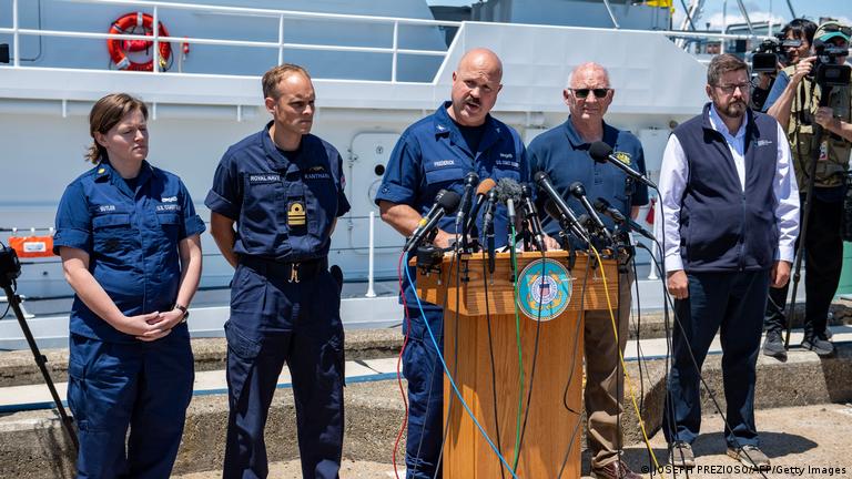 El jefe de la Guardia Costera estadounidense habla en puerto desde un atril repleto de micrófonos con una embarcación de rescate detrás.