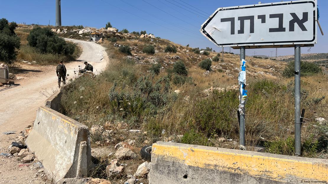 Eine sandige Straße führt auf einen von Büschen bewachsenen Hügel; zwei Menschen sitzen auf Betonbarrieren; ein Schild mit hebräischer Beschriftung weist die Straße hinauf