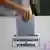 Se ve la mano de la electora metiendo su papeleta en una urna en la que se lee "presidente y vicepresidente".