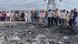 A small crowd of people is looking down a damaged and blackened piece of road with the overturned bus behind them A small crowd of people is looking down a damaged and blackened piece of road with the overturned bus behind them