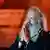 Rick Rubin, a man with a long grey beard, holds his hands in thankful prayer on stage in front of a mic. 