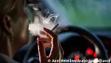 Eine Frau sitzt mit einer brennenden Zigarette hinter dem Lenkrad ihres Autos.