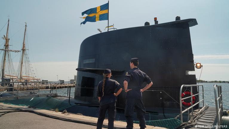 The top of a submarine with a Swedish flag flying from it, alongside a dock with masts of sailing ships in the background. A man and a woman in dark blue uniform stand with their backs to the camera.