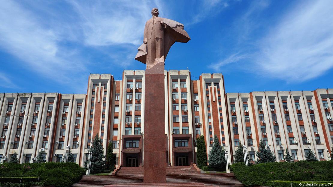 Una estatua de Lenin con el abrigo ondeando, fotografiada desde abajo, se alza sobre un alto pedestal frente a un edificio amplio y representativo.