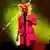 A cantora e compositora brasileira Bia Ferreira, em foto da cintura para cima, canta em um palco com a mão esquerda sobre o peito e de olhos fechados. Ela é uma mulher negra e usa dreads cor de algodão rosa. Está trajando um sobretudo rosa choque. Por trás dela há uma luz amarela.