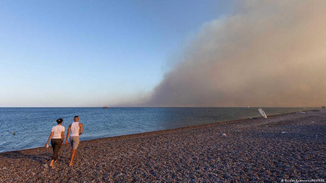 Incendio forestal en Rodos, Grecia, es visto desde la costa. Por la playa caminan dos personas.