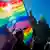 Uma pessoa participando de uma marcha LGBT carrega uma bandeira nas cores do arco-iris. Ao fundo, vê-se as cúpulas douradas de uma igreja ortodoxa