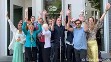 The Innoklusio team can be seen with part of DW's Diversity Management team, standing in front of the entry to the DW building at the Berlin site. They are holding up their arms, waving and smiling.