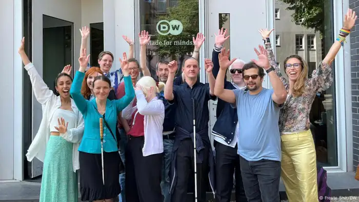 The Innoklusio team can be seen with part of DW's Diversity Management team, standing in front of the entry to the DW building at the Berlin site. They are holding up their arms, waving and smiling.