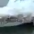 A large white cargo ship in the water. It has a lot of holes in the side and white smoke is billowing out of it.