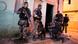 Policiais armados com fuzis em frente a uma casa. Um deles está agachado e inspeciona alguma coisa no chão Policiais armados com fuzis em frente a uma casa. Um deles está agachado e inspeciona alguma coisa no chão