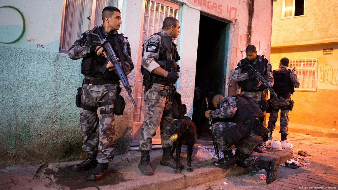 Quatro policiais em pé e um ajoelhado, de costas, ao lado de um cachorro e em frente a uma esquina. 