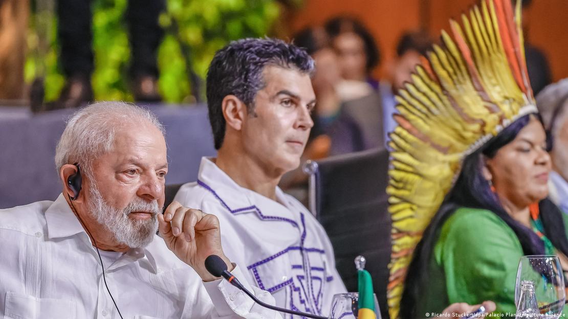 Lula sentado em frente a um microfone. Ao fundo, uma líder indígena usando cocar