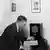 Schwarzweißbild: Ein Mann mit Frisur und Kleidung der 30er Jahre sitzt, nach vorn gebeugt, auf einem Stuhl und dreht am Senderknopf eines Radiogeräts mit der Form und Größe etwa eines hochkant stehenden Schuhkartons