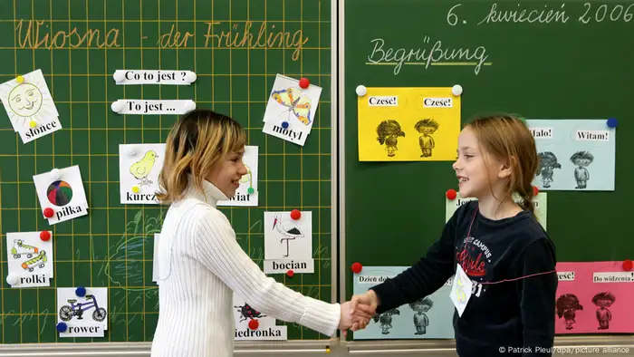 Two girls shaking hands, girl on left in front of a chalkboard with Polish phrases, girl on right with German.