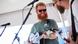 Red-haired and bearded Oliver Anthony smiling and playing guitar.