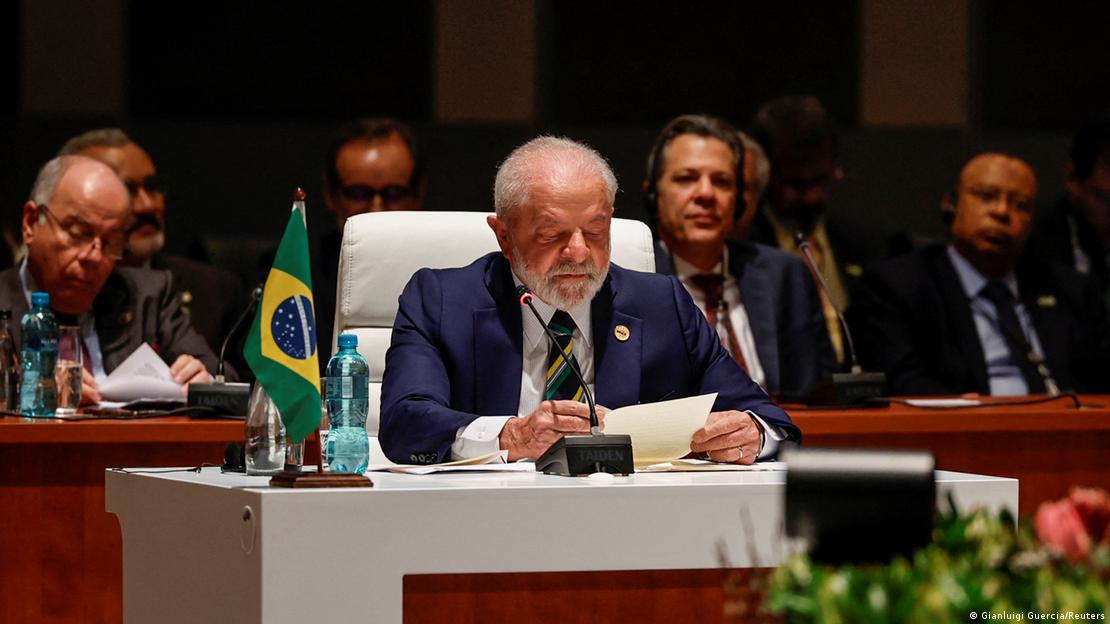 Lula sentado em uma poltrona branca, ao lado de uma pequena bandeira do Brasil que está sobre uma mesa