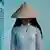 A symbolic image of a young woman in Vietnam, wearing a pointed hat and a white shirt, standing next to blue shutters
