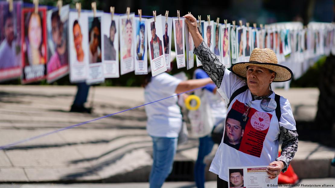 Una mujer lleva una foto de un joven desaparecido y muchas fotos cuelgan en hilera.