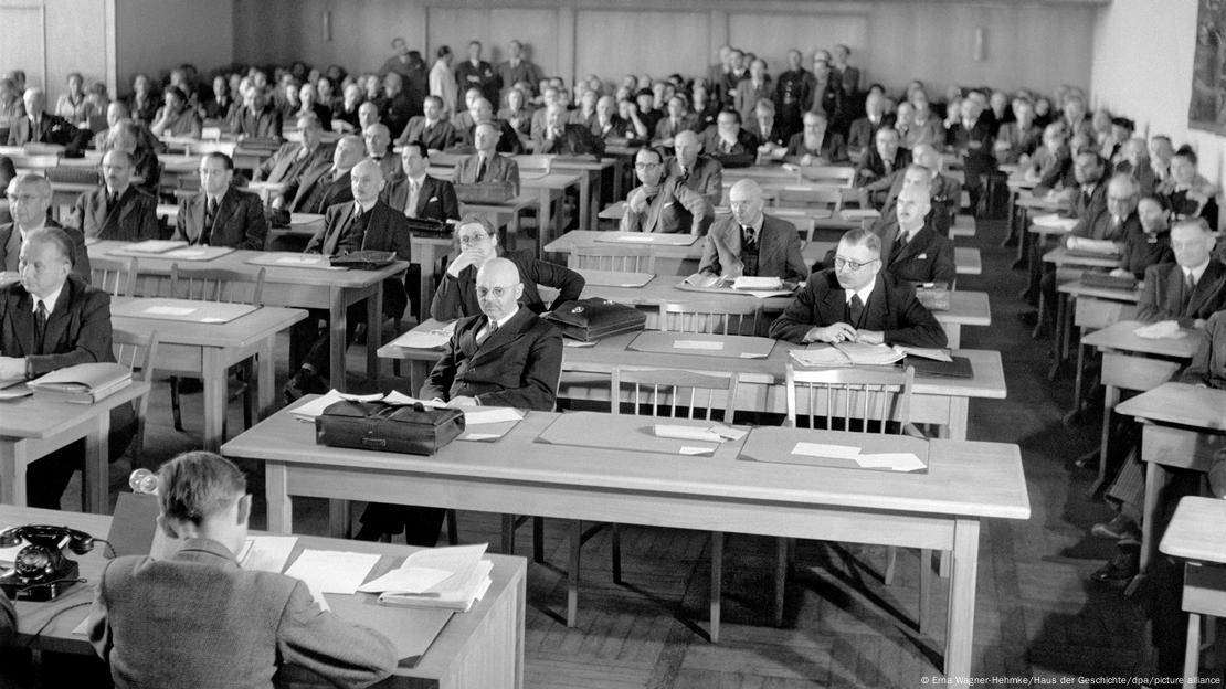 Ausarbeitung des Grundgesetzes vor 75 Jahren in Bonn; historisches Foto. Männer und Frauen sitzen in einer Halle an Tischen und haben Unterlagen vor sich. 