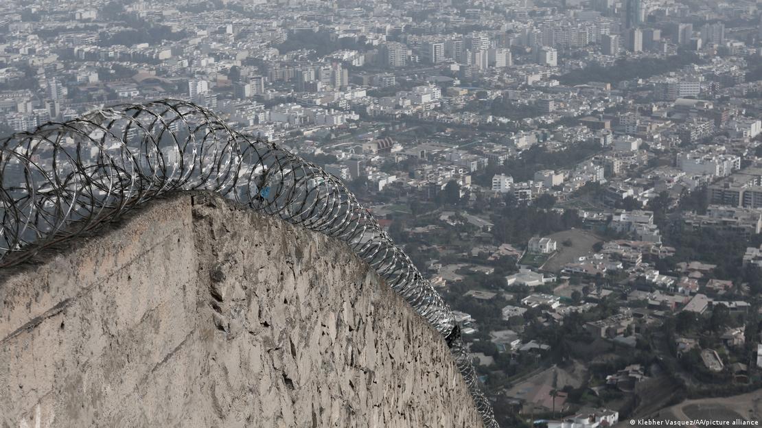 Muro de concreto com arame farpado no topo e vista da cidade ao fundo