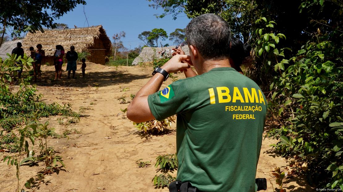 Homem com camisa verde com inscrição IBAMA com cabanas indígenas ao fundo