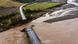 An aerial photograph shows a road across a bridge that has collapsed into a swollen river brown with mud. An aerial photograph shows a road across a bridge that has collapsed into a swollen river brown with mud.