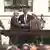 Yitzhak Rabin and Yassir Arafat stand on the lawn at the White House behind a large table. They shake hands as Bill Clinton puts out his arms out behind them