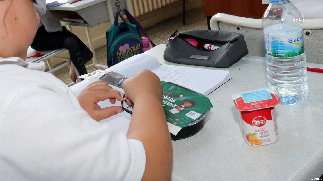 Okulda dağıtılan ücretsiz yemeğinin paketini açan bir çocuğun kolları ve yüzünün bir kısmı görülüyor. 